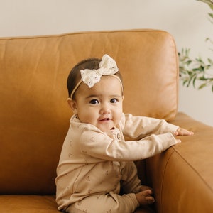 May include: A baby wearing a light brown onesie with a crescent moon and star pattern sits on a brown leather couch. The baby is wearing a white lace bow headband and is smiling at the camera.