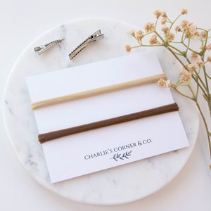 May include: Two nude and brown elastic hair ties on a white card with the text "Charlie's Corner & Co.".
