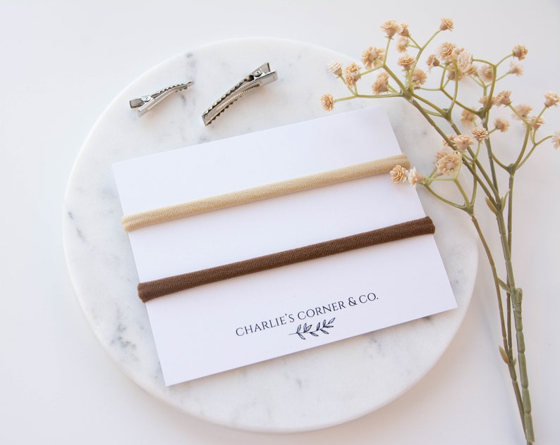 May include: Two nude and brown elastic hair ties on a white card with the text "Charlie's Corner & Co.".