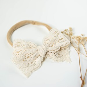 May include: A white lace bow headband with a nude elastic band. The headband is on a white background.