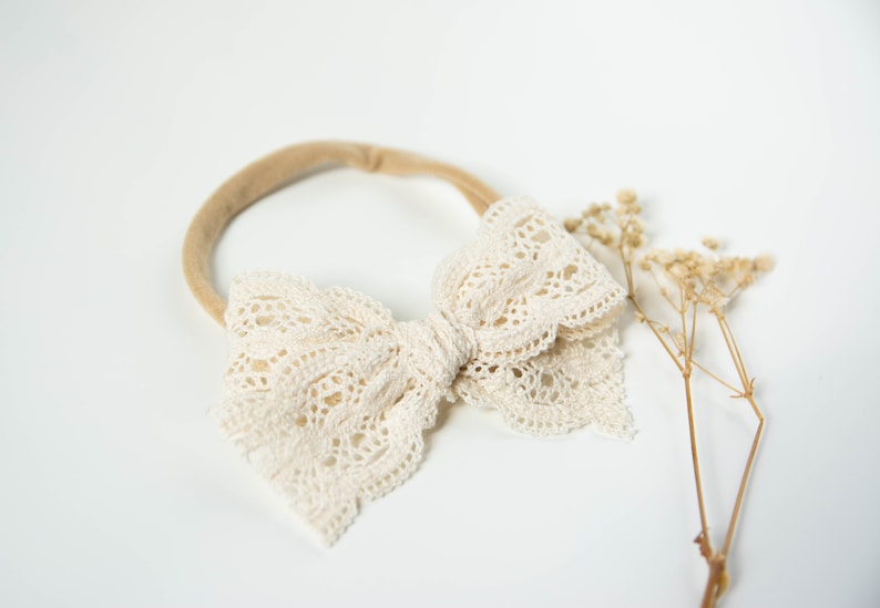 May include: A white lace bow headband with a nude elastic band. The headband is on a white background.