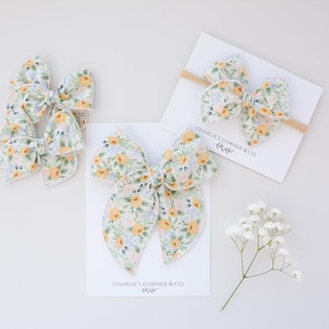 May include: Several floral fabric bows in a variety of sizes are displayed on a white surface. The bows feature a pattern of yellow and orange flowers with green leaves on a white background. One bow is attached to a headband. The text "Charlie's Corner & Co" is printed on a card.