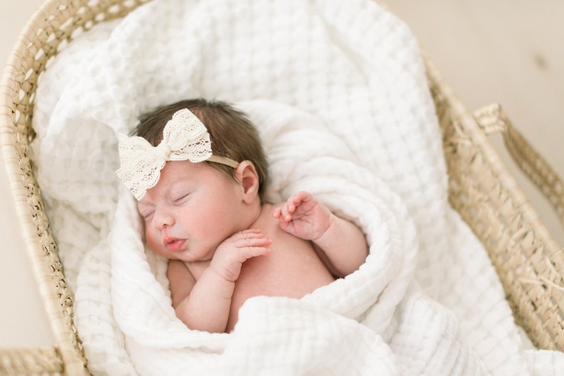 May include: A newborn baby girl sleeping in a white woven basket, wrapped in a white blanket with a white lace bow headband.