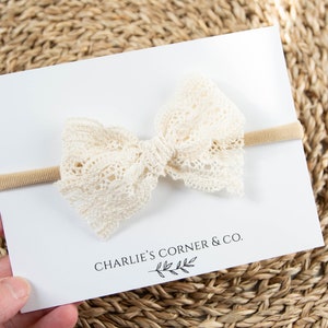 May include: A white lace bow headband with a nude elastic band. The headband is on a white card with the text "CHARLIE'S CORNER & CO."