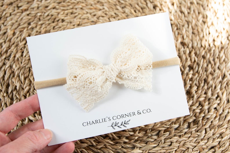 May include: A white lace bow headband with a nude elastic band. The headband is on a white card with the text "CHARLIE'S CORNER & CO."
