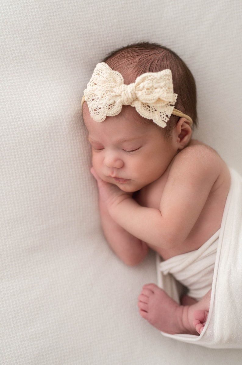 May include: A newborn baby girl sleeping peacefully on a white blanket. She is wearing a white crocheted headband with a bow. The baby is swaddled in a white blanket.
