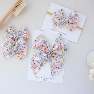 May include: Floral fabric hair bows in pink, yellow, and blue. One bow is attached to a headband, and two are on clips. A wooden brush is in the background. The bows are on white cards with the text "Charlie's Corner & Co."