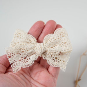 May include: A white lace bow with a scalloped edge. The bow is tied in a knot and is held in a hand.