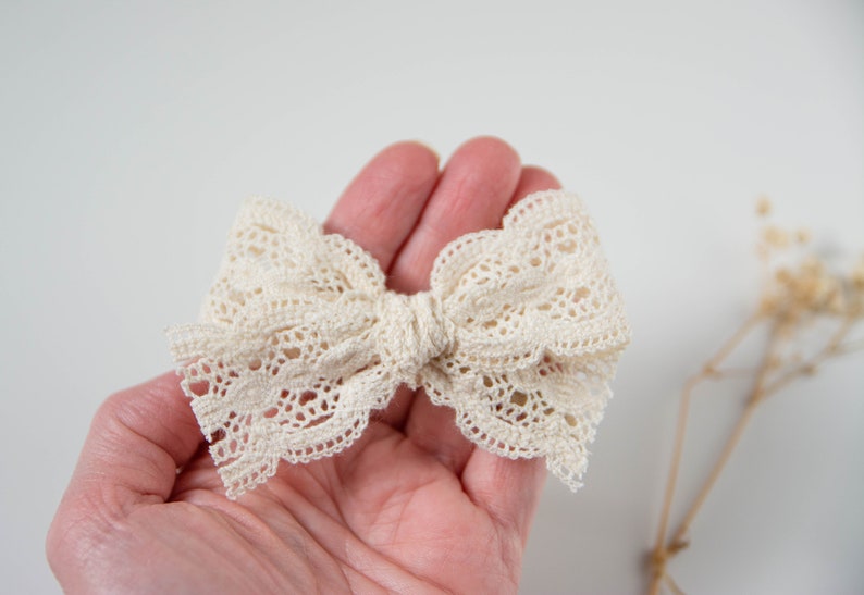 May include: A white lace bow with a scalloped edge. The bow is tied in a knot and is held in a hand.
