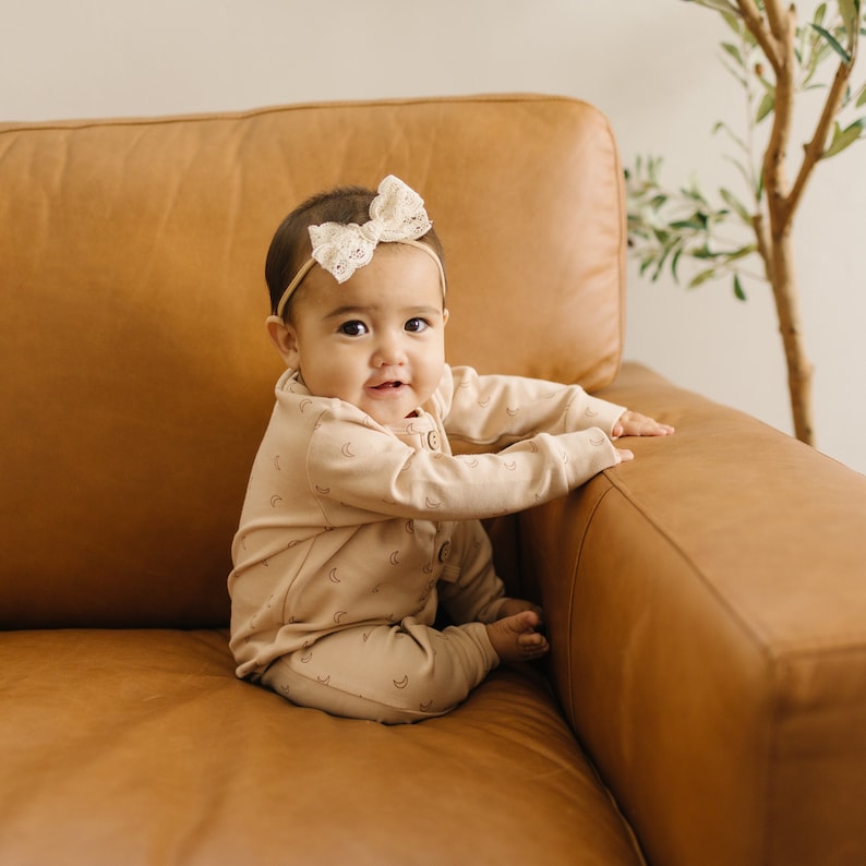 May include: A baby wearing a light brown onesie with a crescent moon and star pattern sits on a brown leather couch. The baby is wearing a white lace bow headband and is smiling at the camera.