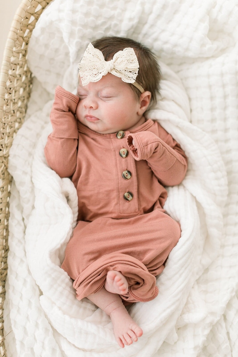 May include: A sleeping baby wearing a  mauve knotted gown with wooden buttons and a white lace headband. The baby is lying on a white blanket in a woven basket.