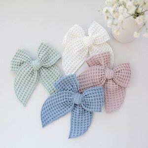 May include: Five waffle-textured hair bows in pastel colors: mint green, white, dusty rose, and light blue. The bows are arranged on a white surface, with a small vase of white flowers in the background.