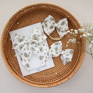 May include: A set of four white fabric hair bows with a floral pattern in blue, brown, and green. The bows are arranged on a woven wicker tray with a white card that reads "CHARLIE'S CORNER & CO."