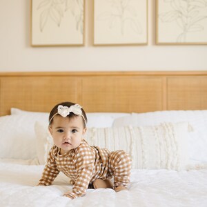 May include: A baby wearing a brown and white gingham onesie with a white bow headband is crawling on a white bed. The bed has a white pillow and a white throw blanket.