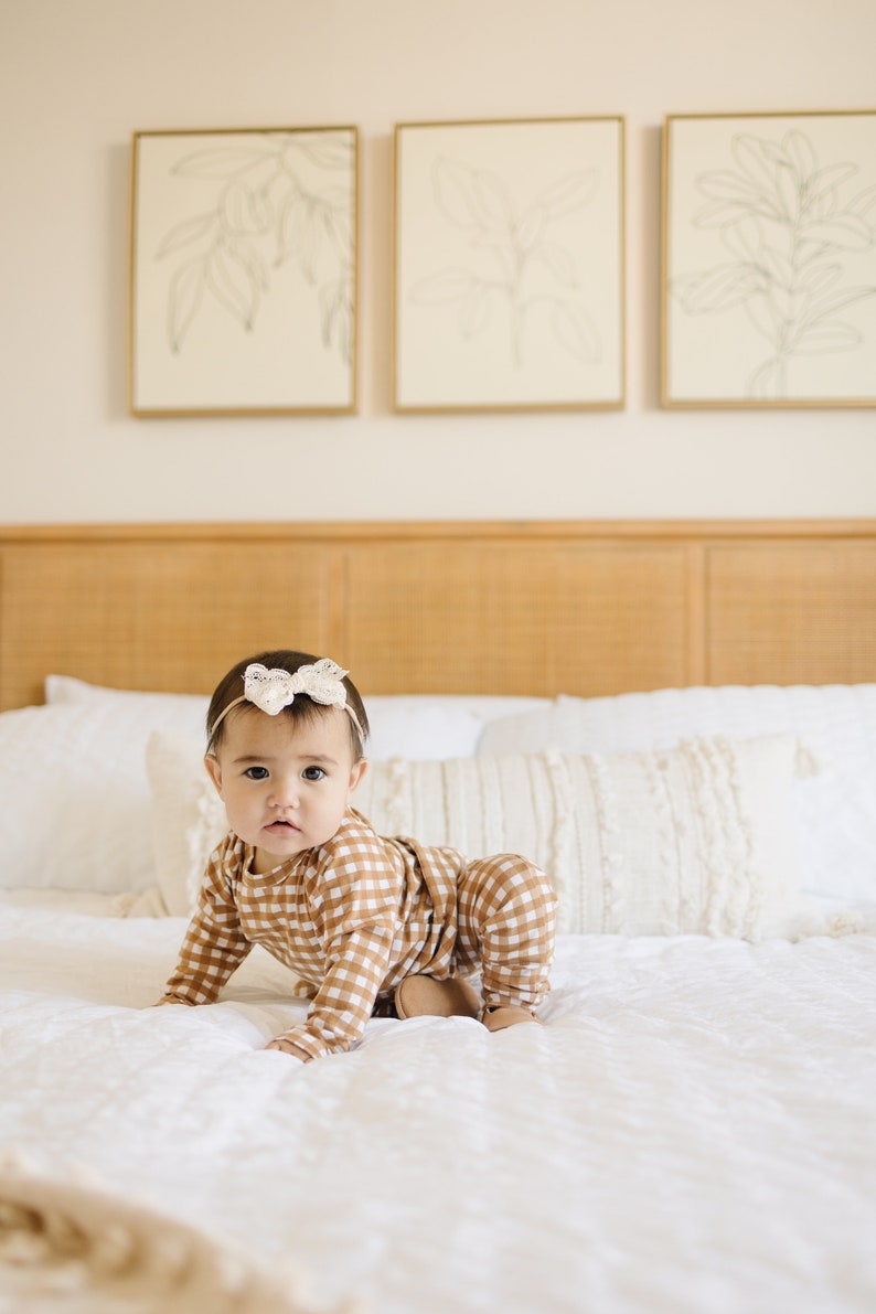 May include: A baby wearing a brown and white gingham onesie with a white bow headband is crawling on a white bed. The bed has a white pillow and a white throw blanket.