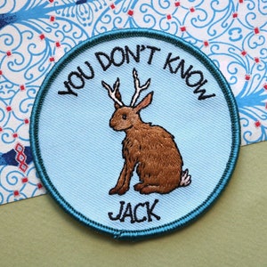 Puede incluir: Un parche bordado azul y verde con un conejo marrón con cuernos. El texto en el parche dice "You don't know Jack".