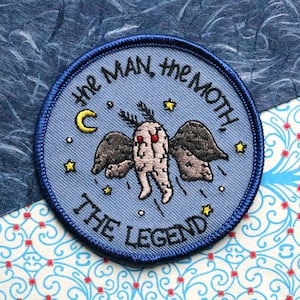 May include: A blue embroidered patch with a white crescent moon, stars, and a moth with a human-like body and wings. The text "The Man, The Moth, The Legend" is written in a circle around the moth.