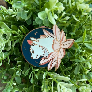 May include: A white cat with blue eyes is depicted in profile, looking to the left. The cat is surrounded by pink and white flowers, and the background is a dark blue circle with gold stars.