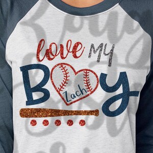 May include: A white and blue baseball raglan t-shirt with the text "love my boy" in red and silver glitter. A red and white baseball heart with the name "Zach" is in the center of the shirt. A brown baseball bat is below the heart.