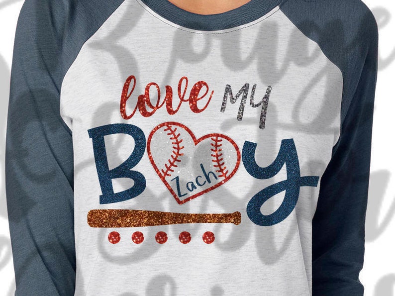 May include: A white and blue baseball raglan t-shirt with the text "love my boy" in red and silver glitter. A red and white baseball heart with the name "Zach" is in the center of the shirt. A brown baseball bat is below the heart.