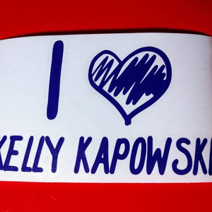 May include: A white sticker with blue lettering that reads "I love Kelly Kapowski". The "love" is represented by a blue hand-drawn heart.