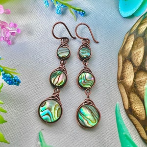 May include: A pair of long, dangling earrings with three iridescent abalone shell beads on each earring. The beads are wrapped in copper wire and the earrings have copper hooks.