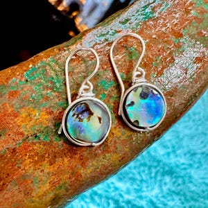 May include: A pair of silver dangle earrings with iridescent abalone shell round pendants. The earrings are on a rustic brown surface.