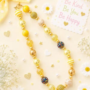 May include: A beaded necklace featuring a variety of yellow, gold, and multi-colored rhinestone spheres, interspersed with smaller gold-tone spacer beads. The necklace includes a toggle clasp and is arranged in a circular shape on a light surface.