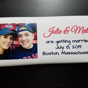 May include: A white chocolate bar with a photo of a couple in a baseball stadium. The text on the bar reads "Julie & Matt are getting married! July 8, 2019 Boston, Massachusetts".