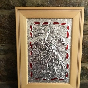 Puede incluir: Una obra de arte de metal enmarcada que representa dos grullas con ojos verdes. Las grullas están en acabado plateado y se colocan sobre un fondo rojo. El marco está hecho de madera y tiene un acabado natural.