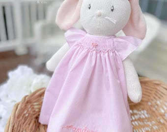 May include: A white knitted bunny doll with pink ears and a pink gingham dress. The dress has a small bow and the name "Anderson" embroidered in pink. The doll is sitting in a woven basket. The text "three little birds & co." is visible.