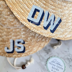 May include: Two woven straw sun hats featuring a white rope chin strap with a dark brown bead. One hat displays the navy blue and light gray letters DW, while the other shows JS. A circular tag reads Three Little Birds & Co. with a bird icon.