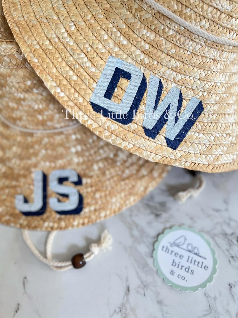 May include: Two woven straw sun hats featuring a white rope chin strap with a dark brown bead. One hat displays the navy blue and light gray letters DW, while the other shows JS. A circular tag reads Three Little Birds & Co. with a bird icon.