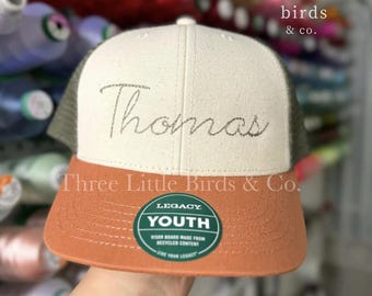 May include: A youth-sized baseball cap with a cream-colored front panel embroidered with the name "Thomas" in a script font. The brim is orange, and the mesh back is olive green. The cap has a "Legacy Youth" sticker.