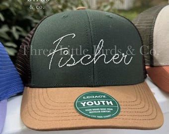 May include: A green and tan youth trucker hat with the name "Fischer" embroidered in white script. The hat has a brown mesh back and a curved tan brim. The hat has a "Legacy Youth" sticker.