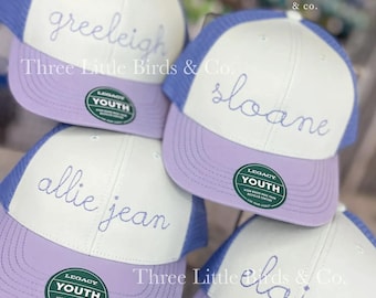 May include: Four youth baseball caps with white fronts, lavender brims, and blue mesh backs. Each cap has a different name embroidered in blue script: Greeleigh, Sloane, Allie Jean, and Elaine. The caps are from Three Little Birds & Co.