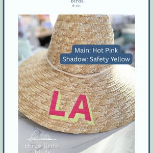 May include: A woven straw hat featuring the letters LA in hot pink with a safety yellow shadow. A white cord encircles the base of the crown. Text on the image reads: Main: Hot Pink, Shadow: Safety Yellow, and the logo for three little birds & co.