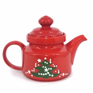 May include: A red ceramic teapot with a lid and handle. The teapot features a green Christmas tree design with white star and dot accents. The teapot is designed for brewing tea and is a festive holiday item.