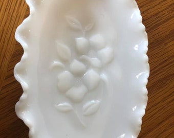 May include: A white, oval-shaped dish with a ruffled edge. The dish features a raised floral design in the center. The dish is made of a glossy, opaque material and is set against a wooden surface.