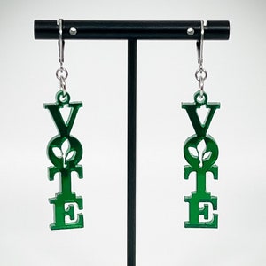 May include: Green acrylic earrings shaped like the word "VOTE" with a small leaf design inside the "O". The earrings are hanging from silver hooks on a black earring stand.