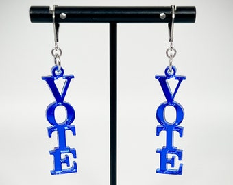 以下が含まれることがあります： 青いアクリル製のイヤリングのペア。各イヤリングには「VOTE」という単語が縦に書かれています。イヤリングは、黒いイヤリングスタンドの銀色のフックにぶら下がっています。