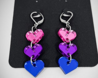 Puede incluir: Un par de pendientes colgantes con tres corazones superpuestos. Los corazones son rosa, morado y azul, y están conectados por anillos plateados. Los pendientes tienen ganchos plateados.