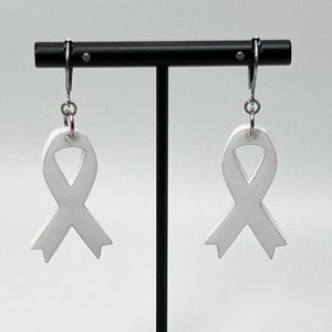 Pode incluir: Um par de brincos de fita branca pendurados em um suporte preto. Os brincos são feitos de um material branco e têm a forma de fitas. Os brincos estão pendurados em ganchos prateados.