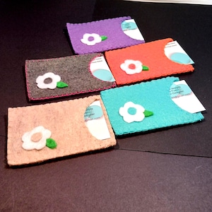 May include: Five felt card holders with a white flower design and a clear plastic window for business cards. The card holders are in different colors: purple, orange, gray, turquoise, and peach.