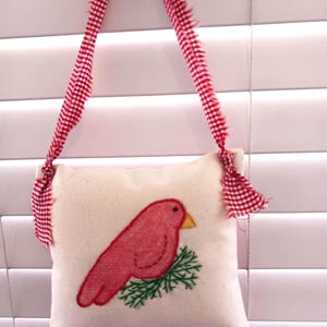 May include: A small, square, off-white pillow with a red bird and green branch embroidery. The pillow is suspended by a red and white checkered ribbon. The bird has a yellow beak.