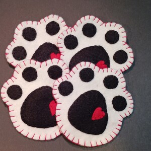 May include: Four white felt paw-shaped coasters with black paw pads and red heart accents. Each coaster is outlined with red stitching. The coasters are arranged in a slightly overlapping pattern.