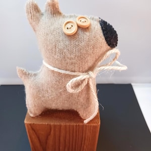 May include: A handmade, light brown knitted dog figurine with button eyes and a black nose. The dog is tied with a white string and sits atop a small, square wooden block. A charming, rustic decorative item.