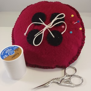 May include: A red heart-shaped pincushion with black felt buttons and white string tied in a bow. The pincushion is surrounded by sewing pins with colorful heads. A spool of white thread and a pair of small silver scissors are also in the image.