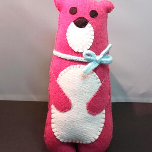 May include: A handmade pink felt otter plush toy with a light blue bow. The otter has a white belly and face, with brown eyes and nose. The plush is approximately 10 inches tall.
