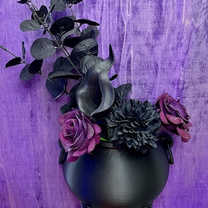 May include: A black cauldron-shaped wall planter filled with dark purple and black flowers and foliage. The planter is mounted on a purple wooden surface. The arrangement includes roses, calla lilies, and other decorative elements.